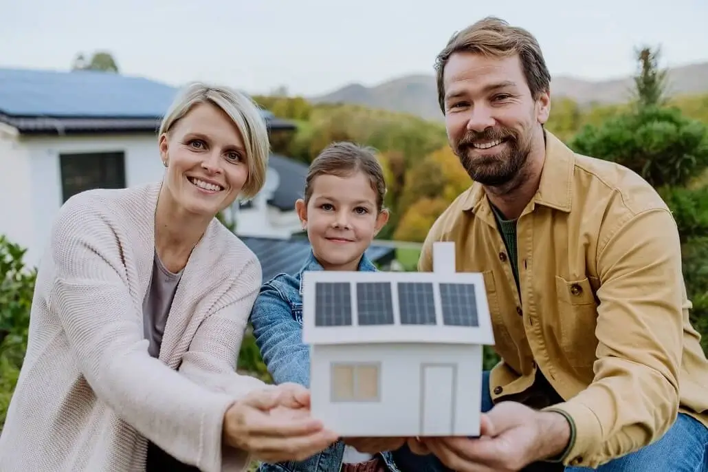 Fröhliche Familie steht vor ihrem Haus und zeigt ein Modell eines Hauses mit Photovoltaik-Anlage